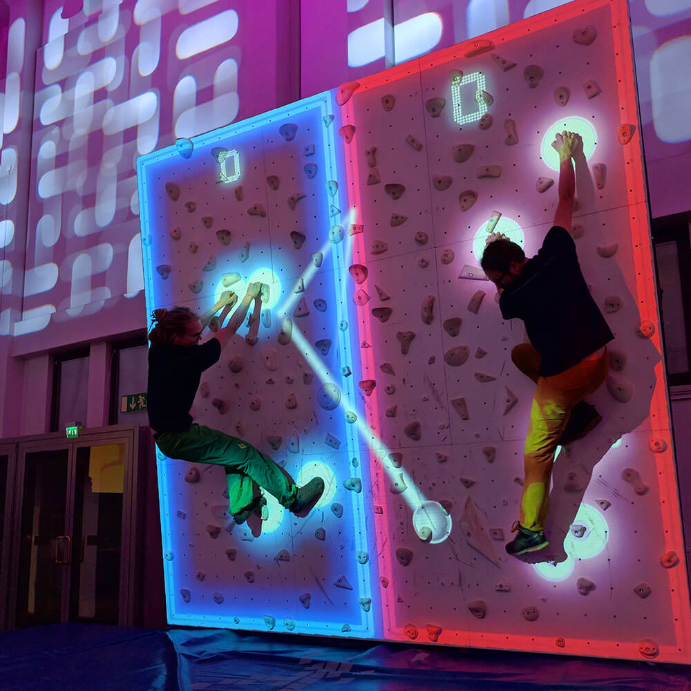An engaging interactive climbing wall where participants scale colorful holds while dynamic projections respond to their movements. The wall features vibrant pink and blue lighting effects that create trails and visual feedback, turning physical activity into an immersive digital playground experience.